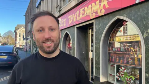 Kyle Luscombe is wearing a black crewneck top and standing in front of his Sweet Dylemma shop