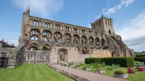 HES A view of Jedburgh Abbey - an impressive old building with towers and a pathway leading up to it through grassy lawns