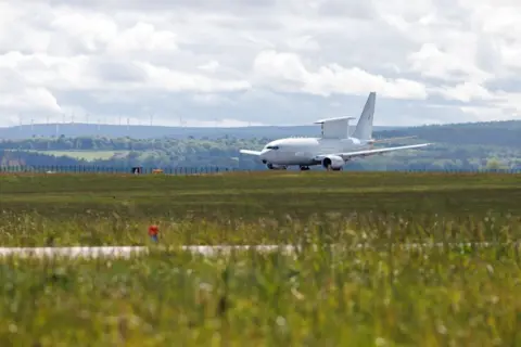 MOD: Crown Copyright Wedgetail airborne early warning (AEW) plane on a runway, there are wind turbines on a hillside in the distance under a cloudy sky.