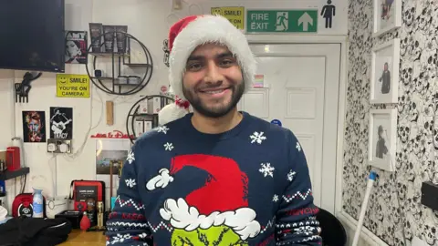Umar Ali is stood inside the barber shop and is wearing a Grinch Christmas jumper and a red Santa hat. Behind him are pictures of the wall and posters of musicians, amongst various hair products. 