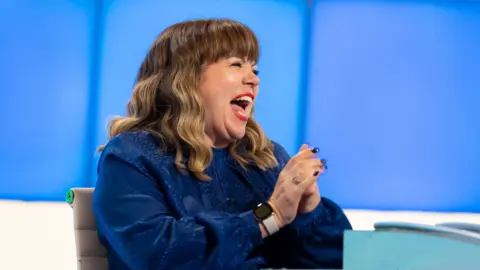 Zeppotron, an Endemol Shine Company Briony May, on the set of the TV show Would I Lie To You, sitting behind a desk, with her hands clasped. She is wearing a shiny dark blue shirt and is laughing.