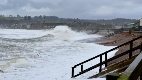Jimmicks/BBC Weather Watchers Large waves crashing on the shore at Stonehaven. The waves are frothy and white. There are rows of houses and other buildings lined along the shore.