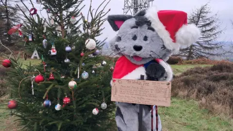 Shropshire Cat Rescue A large grey cat mascot in a santa costume, standing behind a brown sign and a green Christmas tree with baubles hanging from it