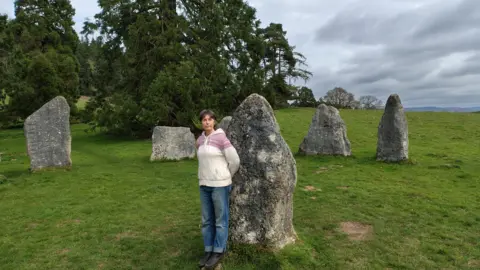 Simon Furber/BBC Eleanor Anstruther, in a white jumper and blue jeans, stands by the rocks.