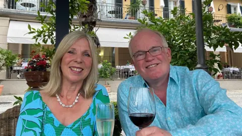 Supplied Jill and Nick Apperley sat next to each other. Jill is holding a glass of white wine and Nick is holding a glass of red wine. Jill is wearing a green and blue top and Nick is wearing a blue shirt. They are both smiling and looking into the camera.