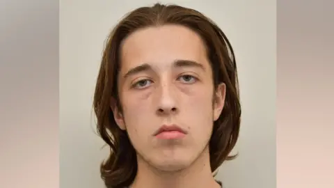 Alfie Coleman has shoulder-length brown hair swept backwards. He looks tired but shows no emotion while pictured in this custody mugshot.