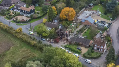 A drone shot of the a destroyed detached house close to other large houses which are untouched. Police tape and emergency vehicles fill the road outside