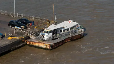 A white ferry with a flat platform around the main body of the boat docks in the sea, with a ramp going up to the dock and two black cars parked nearby.