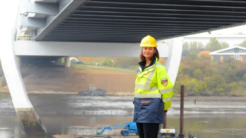 Supplied Amy Dillon has short brown hair. She is wearing a high visibility jacket and a yellow helmet as she stands beneath the Northern Spire bridge next to the banks of the River Wear.