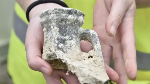 A close-up of someone's hands holding part of a broken Roman-style flaggon with a lipped top and a small handle