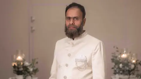 Humayra Ali Masrob wearing white. He has a grey/black beard, and dark hair and is dressed in a formal white jacket. The background shows flower arrangements and candles.