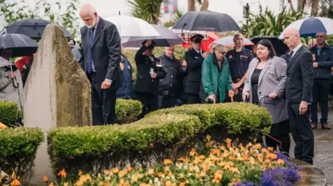 DOUGLAS COUNCIL/MATT MOSUR A man in a suit bows his head while standing in front of the memorial, while is made up of three pillars. A group of other people look on.