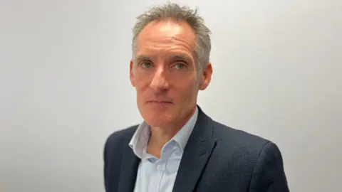 A man wearing a light shirt and a dark blazer. He has short grey hair. He is staring directly at the camera.