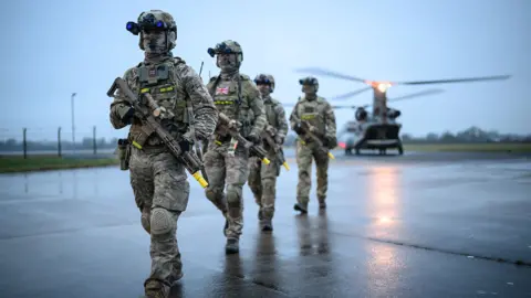 Leon Neal / Getty Images A line of British soldiers walk towards the camera while wearing camouflage uniforms and holding guns on the tarmac at RAF Leeming with a Chinnook helicopter on the ground in the background during a grey foggy morning while training in Catterick on 30 January.