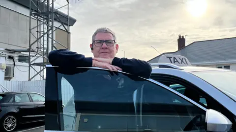 Ian Vaudain, leaning on the door of his taxi. He is wearing glasses and a dark jumper, and has short grey hair.