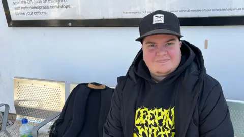 Dean Carter, pictured sitting on a row of metal seats outside the bus station. He is wearing a cap and a thick, hooded coat, over a dark hoodie with a yellow design on the front. His rucksack is placed on the chair next to him.