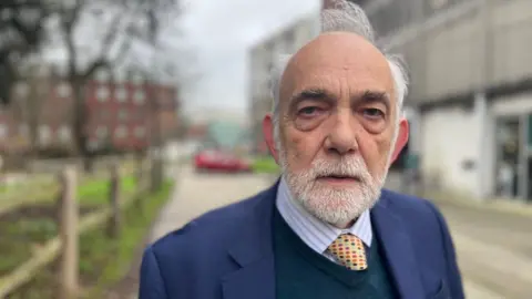 Michael Mitchell in a striped shirt and patterned tie with a navy blue V-neck sewater and a navy blue suit jacket and a blurred background of Dix's Field in Exeter.