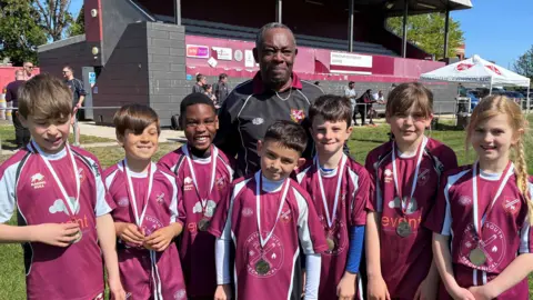 A group of young rugby players wearing maroon Streatham and Croydon RFC kits and medals pose on a grass pitch in front of the club’s stand at Frant Road, with an adult coach standing behind them and other people, buildings and banners visible in the background on a sunny day.