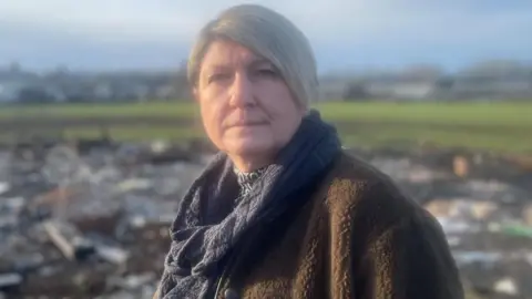 Simon Thake/BBC A woman with short light hair and dressed in a warm brown fleece and silver scarf stands in-front of a pile of charred rubbish spread over a grass field