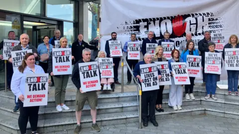 People holding placards stand on the steps of the Strule Arts Centre in a demonstration to raise their concerns about delays to upgrading the A5