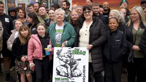 A group shot of men and women stood in front of a mural. They are holding a poster which shows a man sat on rocks, playing a guitar. On the poster are the words Our Billy. Warrior. One Love. You'll never walk along