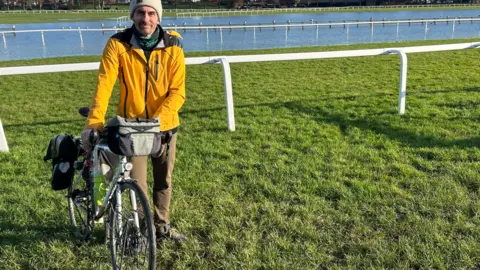 BBC A man wearing a yellow coat and brown trousers is holding a bike on grass and looking at the camera. White railings, water and trees are in the background. 