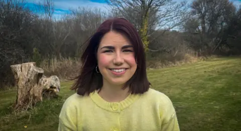 Ffion in a field looking at the camera and smiling. She has a dark shoulder length hair and is wearing a yellow sweater