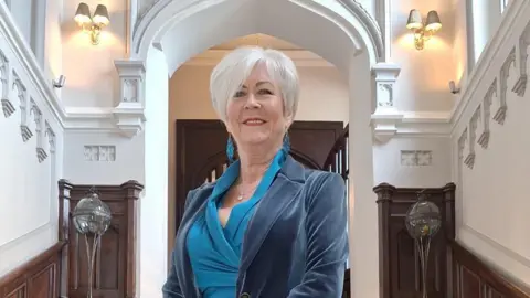 Maggie Montgomery A modern-day image of Maggie Montgomery. She has white hair in a cropped and layered style. She is wearing a teal coloured top with a darker blue velvet blazer over the top. She is standing on a grand dark wood staircase with a white plaster arch in the background. 