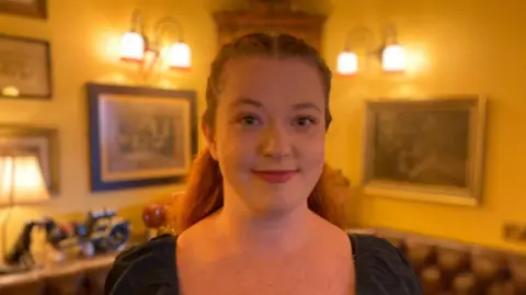 A woman with her hair tied back is standing in what appears to be a pub. There is leather seating behind her and elaborate light fittings on the wall. There are also paintings on the wall behind her, and items on a shelf.