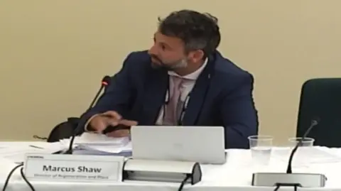 Marcus Shaw appears to be talking into a microphone while sitting at a desk during a council meeting. A sign in front of him includes his name and job title.