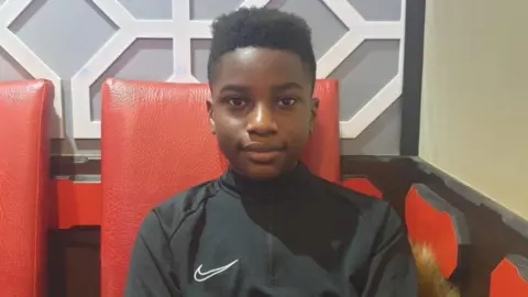 Ashraf Habimana smiling at the camera. He's wearing a black Nike tracksuit and sitting on a red chair