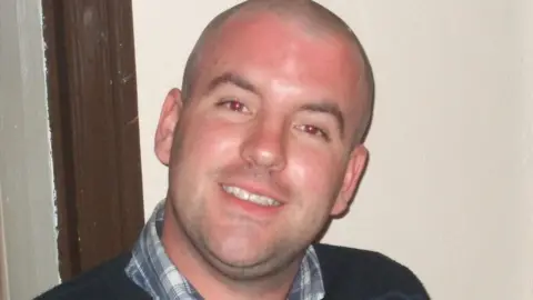 A smiling man wearing a shirt with a dark striped jumper on top