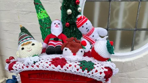 Shirley Thomas Elves dressed in red and green are decoroating a crocheted red, white and grene topper and there appears to be an owl and a Christmas pudding. The festive group have got two Christmas trees behind them, complete with decorations.