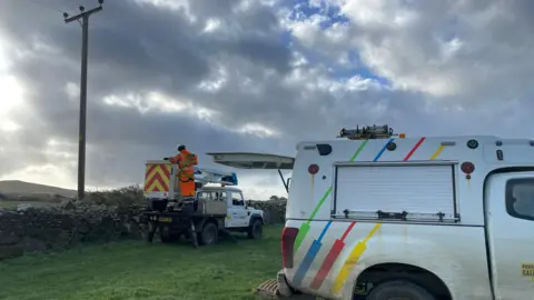 Two National Grid vehicles and an employee in an orange outfit in a field with a wall and power cable overhead.