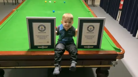 Jude, aged two, who has light brown hair and is wearing a blue t-shirt and black trousers, is sitting on the edge of a snooker table smiling next to two certificates.