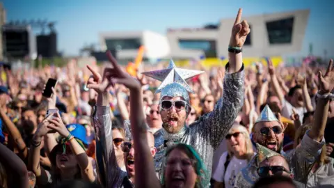 Getty Images Eurovision fans watch the 2023 Eurovision Song Contest final on a giant screen in host city Liverpool. Many fans are dressed up, including the man in the centre of the photo who wears a silver jumpsuit and sunglasses and wears a silver cap with a giant silver star on the top.