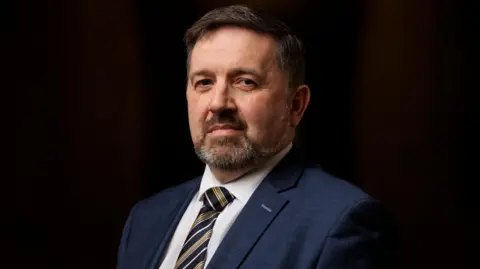 PA Media Robin Swann is looking directly at the camera. He has black hair and a grey beard. He is wearing a white shirt, stripe tie and navy blazer. 