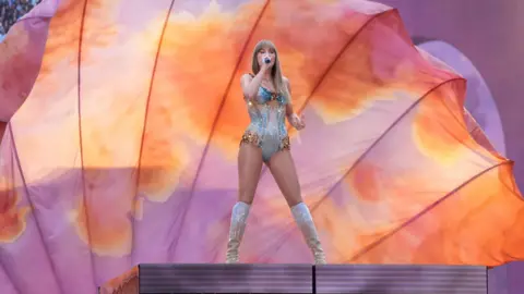 PA Media Taylor Swift on stage at a London concert, in front of a large peach and light purple coloured backdrop. She is singing into a microphone and wearing a one-piece and knee-high boots. 