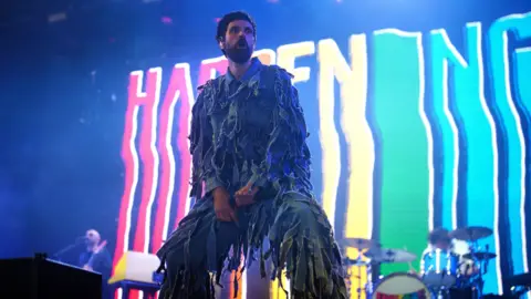 PA Media Serge Pizzorno from rock band Kasabian is pictured performing. He is wearing a blue denim jump suit with shredded pieces of denim hanging from it all over.