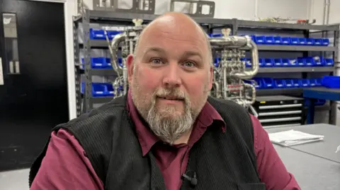 Who are the workers who will fire Scotland's space industry? 3 Derek Harris  fom Skyrora, a bald man with a light coloured bears, wearing a black waistcoat and a burgyndy shirt. He is smiling. Behind him are shelves with rows of blue containers and a work table with a metal structure on it.