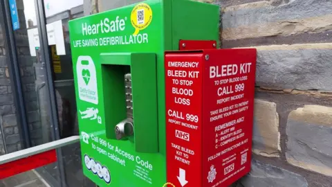 Avon and Somerset Police A green defibrillator on a wall. A red bleed kit box has been installed next to it. It reads "Emergency bleed kit to stop blood loss" with the 999 emergency number and NHS logo underneath in white letters.