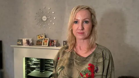 A photo of Amy Geary, with long blonde hair and wearing a green top, standing in front of a fire place 