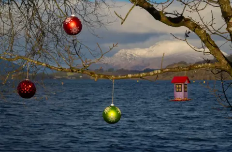 Andy Inglis Christmas tree baubles hanging on branches of a tree with a loch in the background