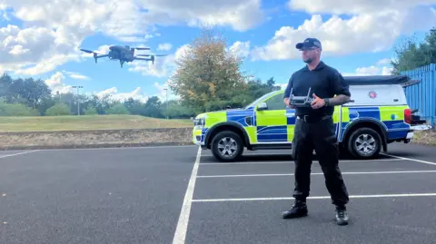 Photograph of PC Tony Ashurst, who is a drone pilot for Greater Manchester Police. The image shows him in his uniform, with the drone in front of him.