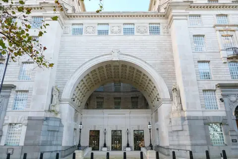 Getty Images The grand stone entrance of Thames House, the London headquarters of MI5, showing a large arched gateway, carved stonework and glass-panelled doors, with black security bollards lining the pavement in front.