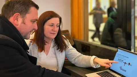 Julia Buckley A dark-haired man in a black coat and and dark-haired woman in a grey blazer and white top, looking at a laptop with the woman pointing at the screen.