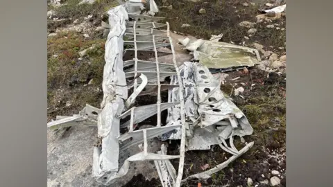 Chris Bullock The remains of part of a World War Two plane, resting on grass and rock. A grid of metal has been revealed, and it is wing-shaped. There is more metal crumpled beneath it.