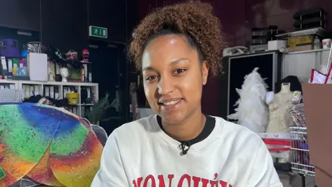 Lily-May Symonds/BBC Chanice Thomas is smiling and wearing a white jumper. Behind her are mannequins and colourful costume materials.