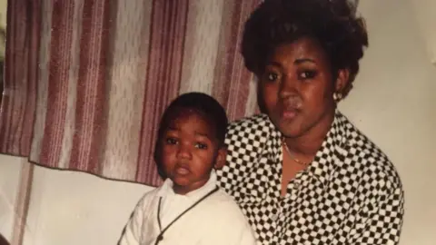 family handout A baby wearing a white cardigan sitting with a women wearing jewellery and a white and black jacket 