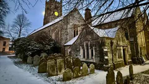 jobear/BBC Weather Watchers A church and small graveyard. There is snow on the ground and on the roofs of the church building. There are trees around. The sky is a light blue.